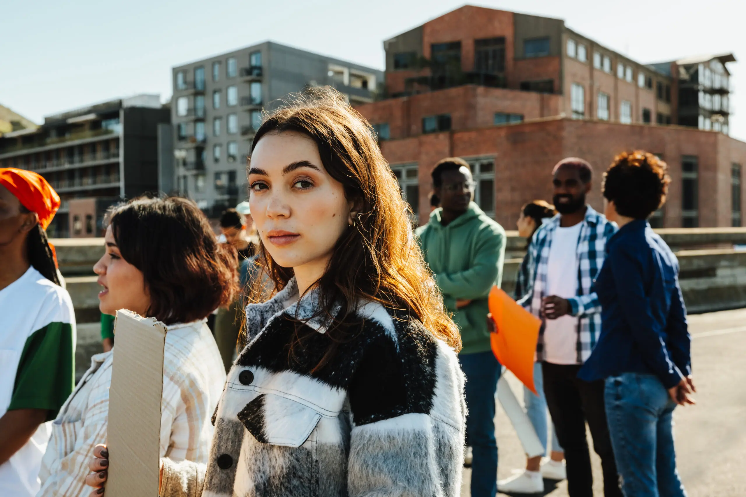 An outdoor gathering of diverse young individuals engaged in social and communal activities during the day. The setting includes an urban background with mixed architecture.