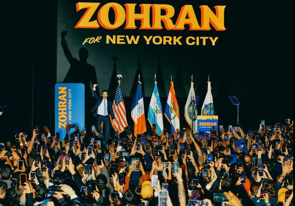 Rally shot in Brooklyn at dusk, crowd behind him blurred, Mamdani stepped forward — hopeful framing of his win